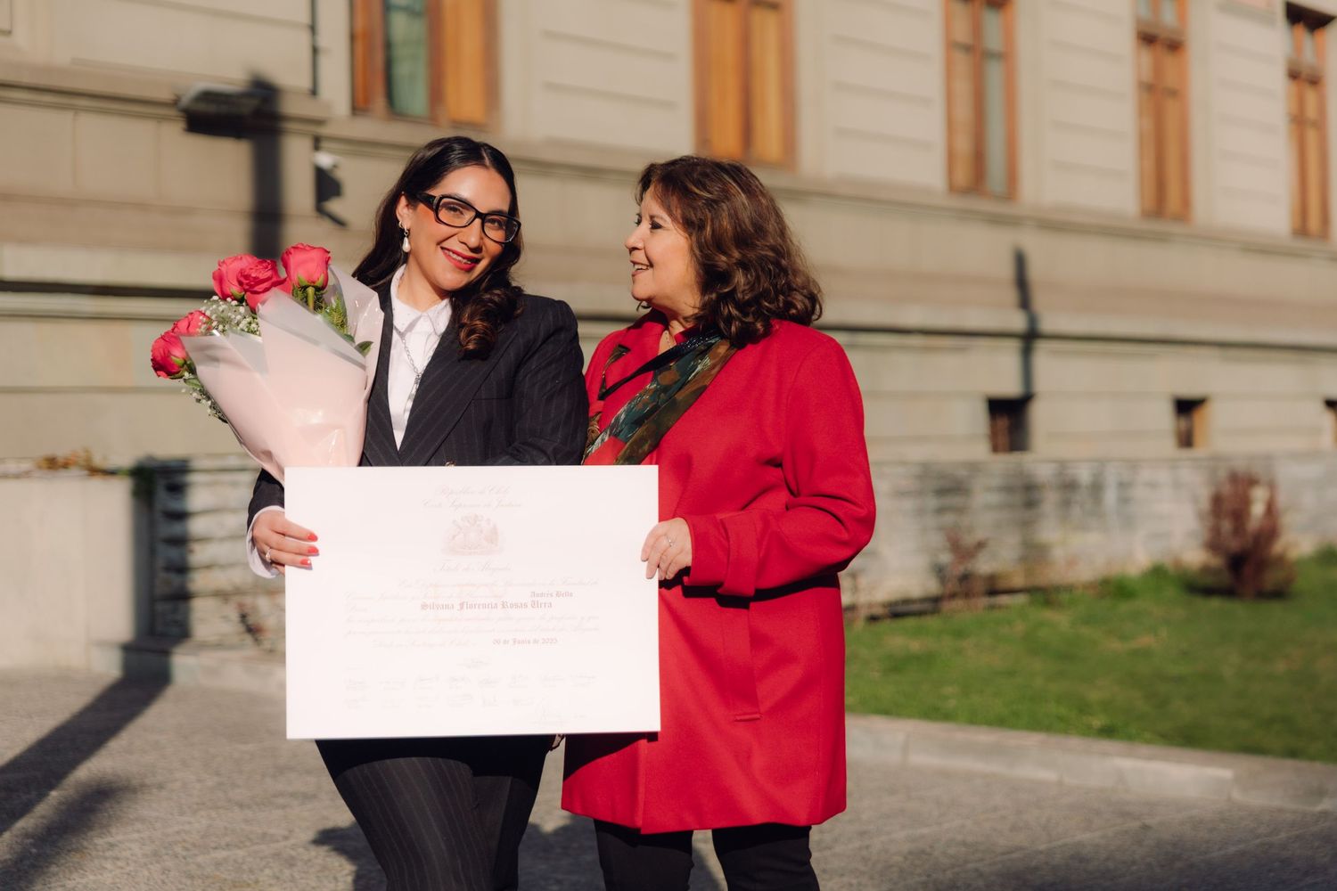Two women sharing joyful graduation moment with diploma and roses outdoors.
