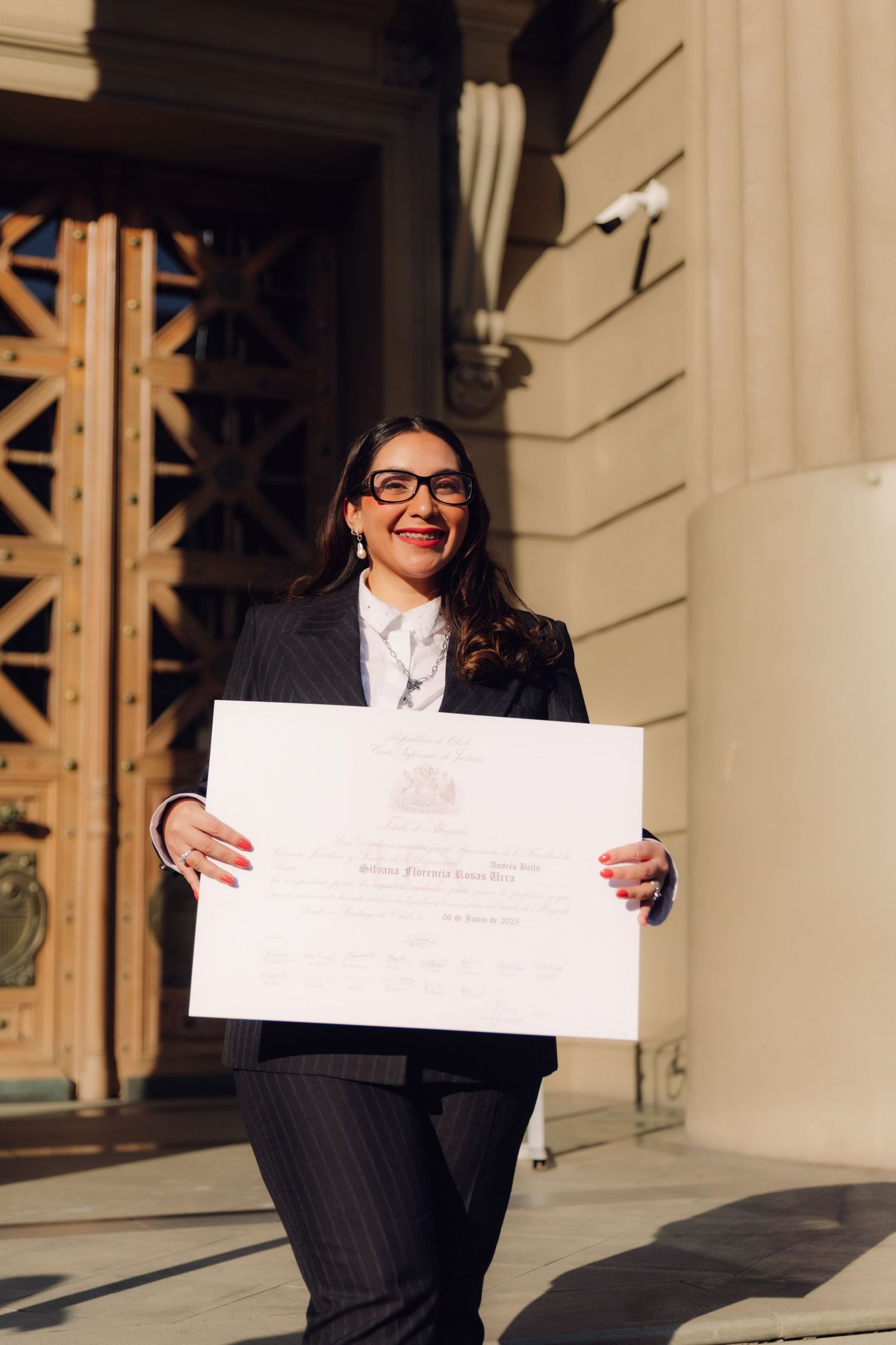 Woman in pinstripe suit proudly displaying diploma at university entrance steps.