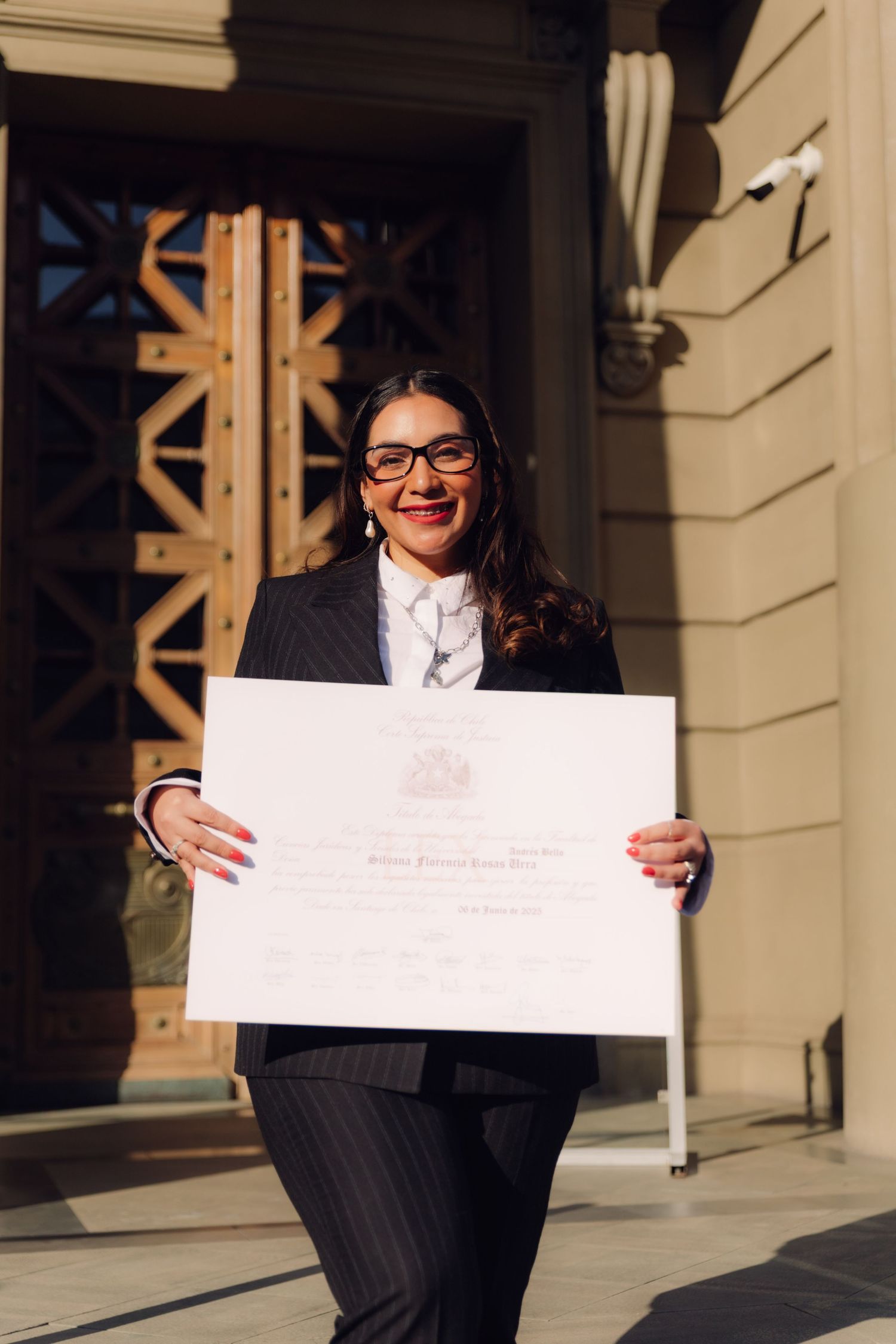 Smiling graduate in black suit holding large diploma outside university entrance.
