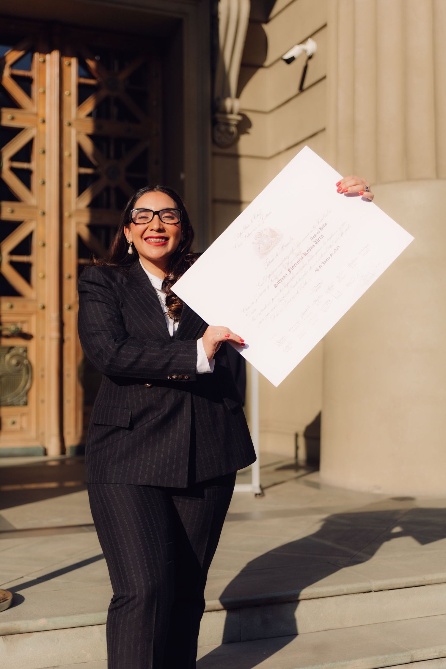 Happy woman in black suit holding diploma aloft outside university building.