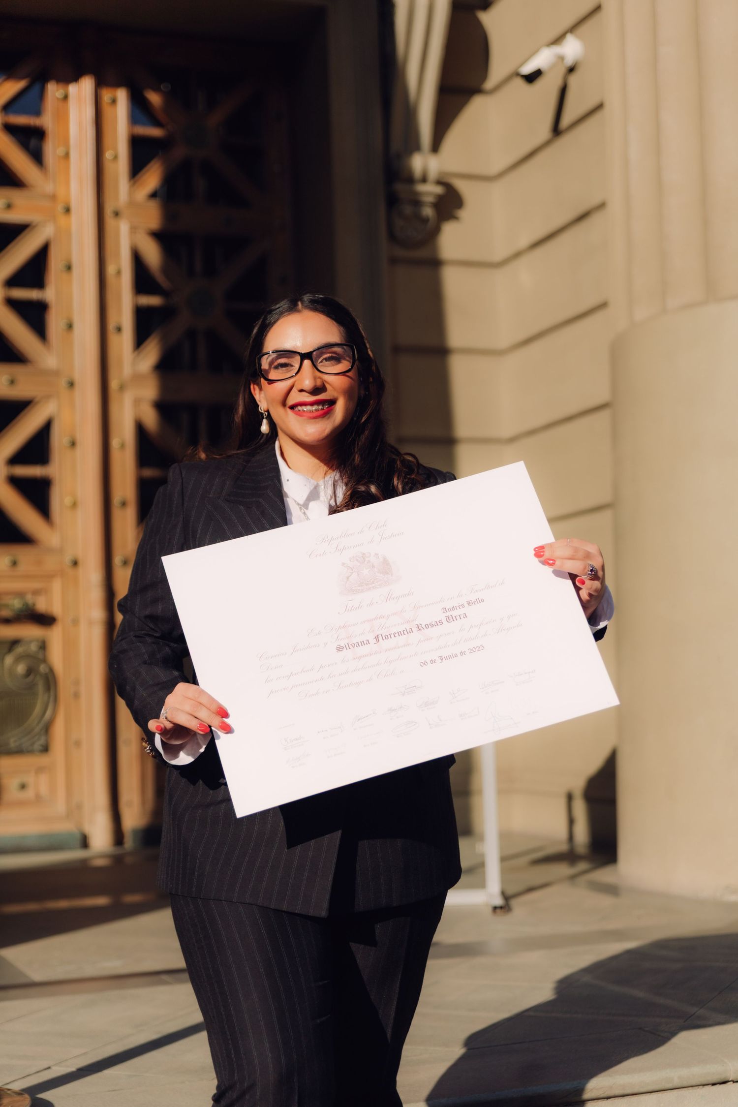 Proud graduate in pinstripe suit displaying official diploma outside university entrance.