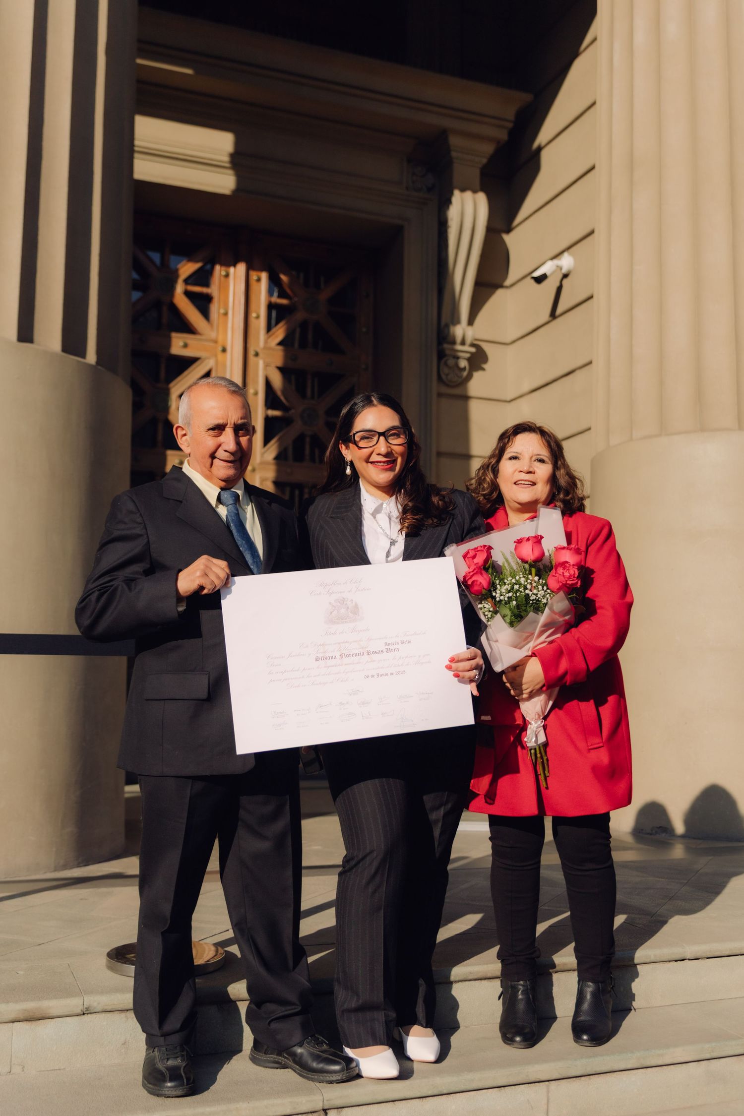Three people pose proudly on building steps holding a large certificate and bouquet of red roses.