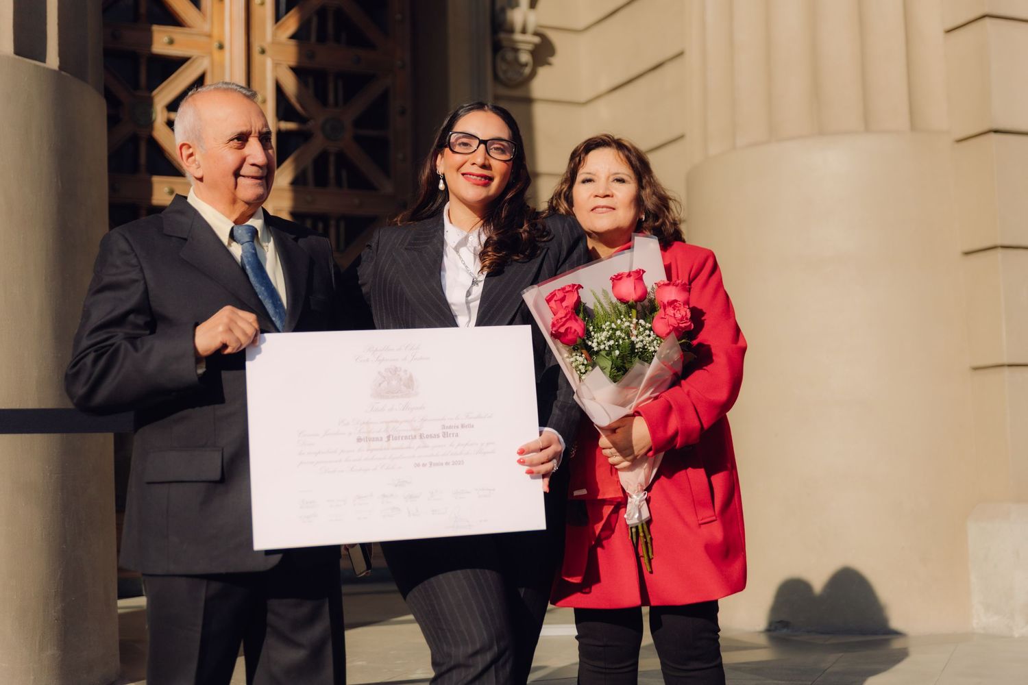 Group of three celebrating outdoors with a framed certificate and red roses bouquet.