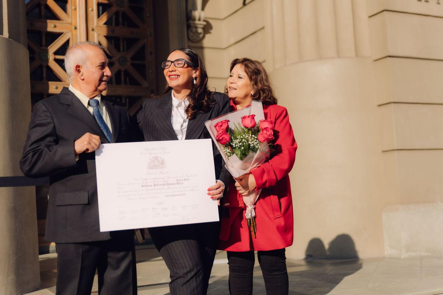 Three people smiling together holding a certificate and red roses outside a classical building.