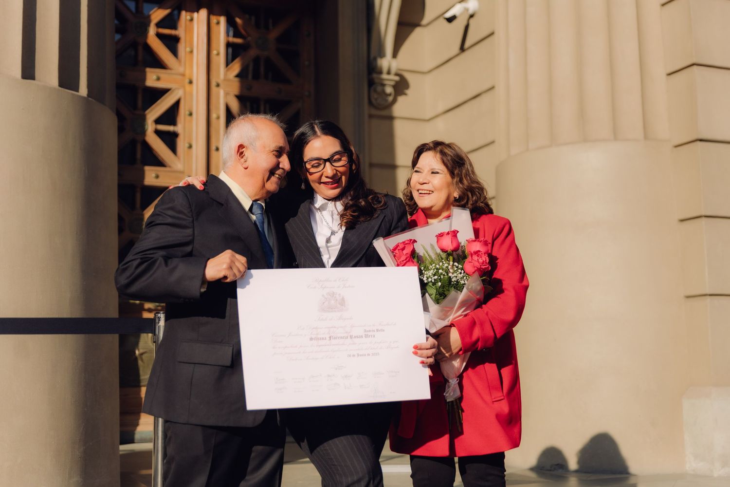 Joyful trio posing with a diploma and red roses outside a formal building entrance.
