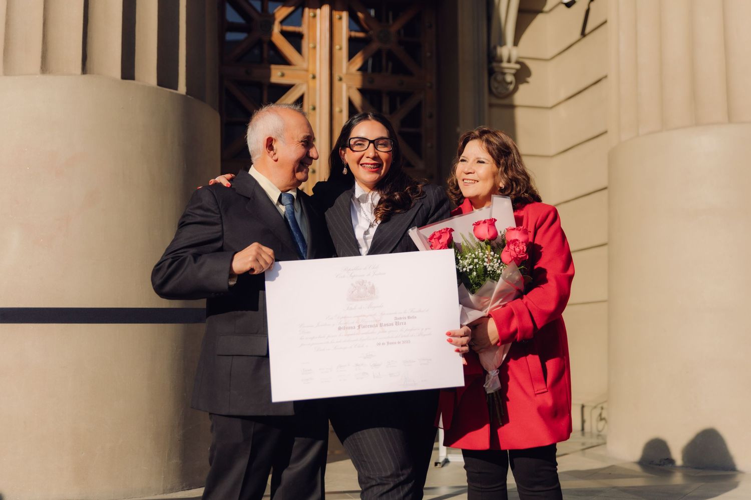 Three people embracing and smiling while holding a graduation certificate and red roses.