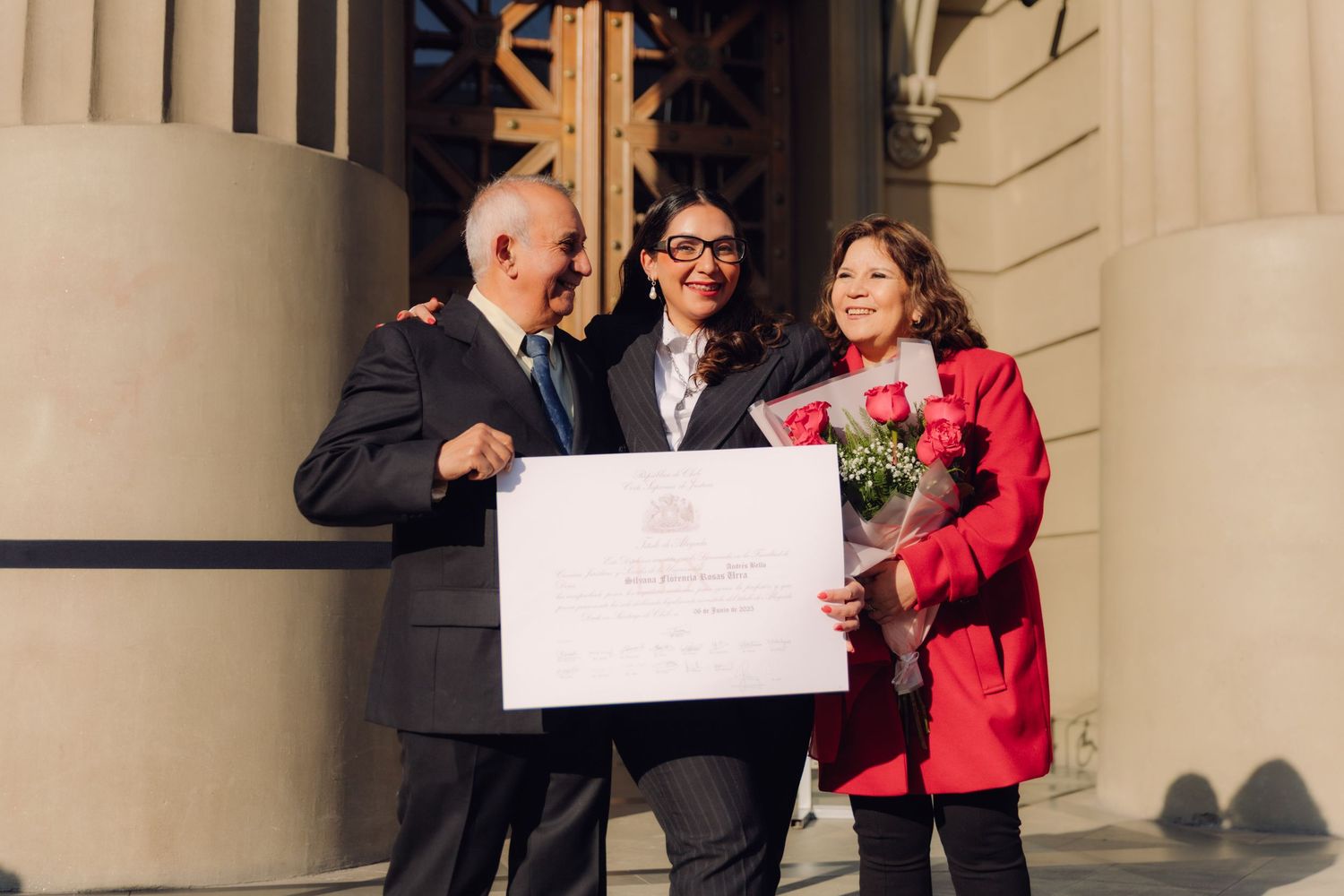 Happy group of three celebrating a graduation with a certificate and red roses outdoors.