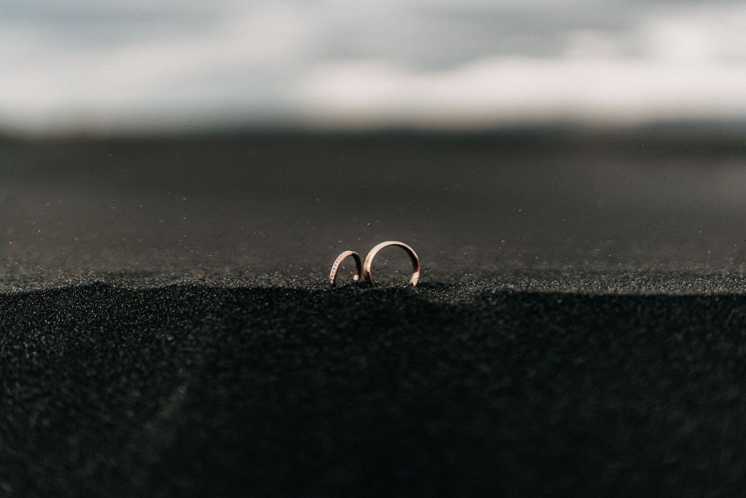 Two gold wedding bands resting in black volcanic sand with ocean blur in background.