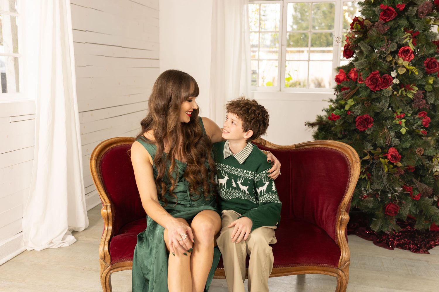 A woman and boy in green holiday outfits sit on a red velvet sofa beside a decorated Christmas tree.