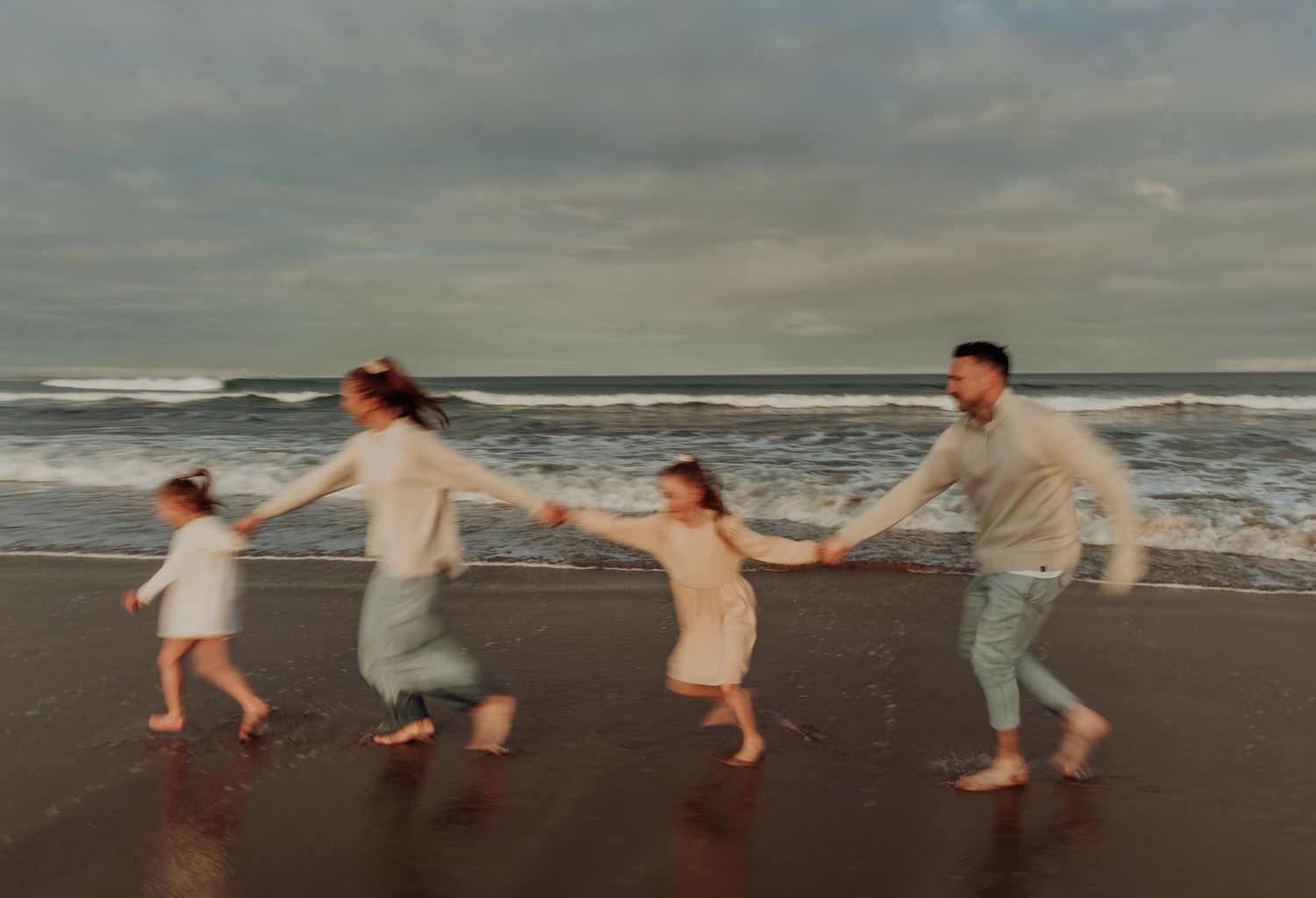 Family of four joyfully running hand-in-hand along a beach with ocean waves in the background.