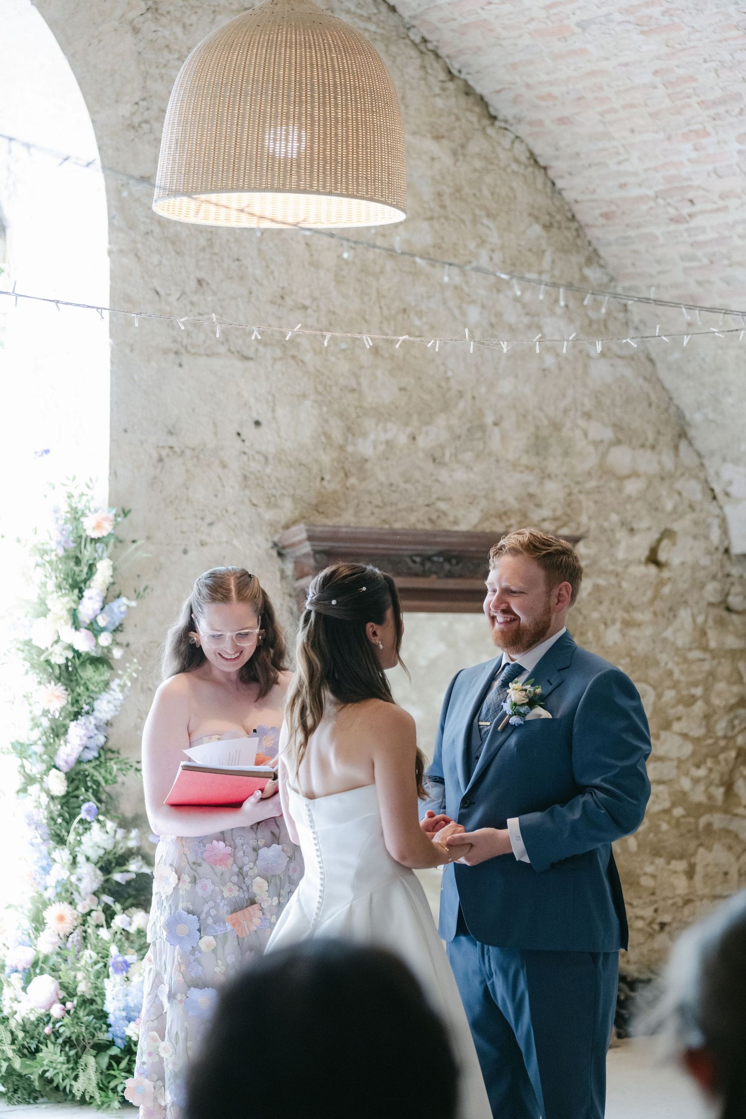 Bride and groom exchange rings during an intimate wedding ceremony in a rustic stone venue with floral decor.