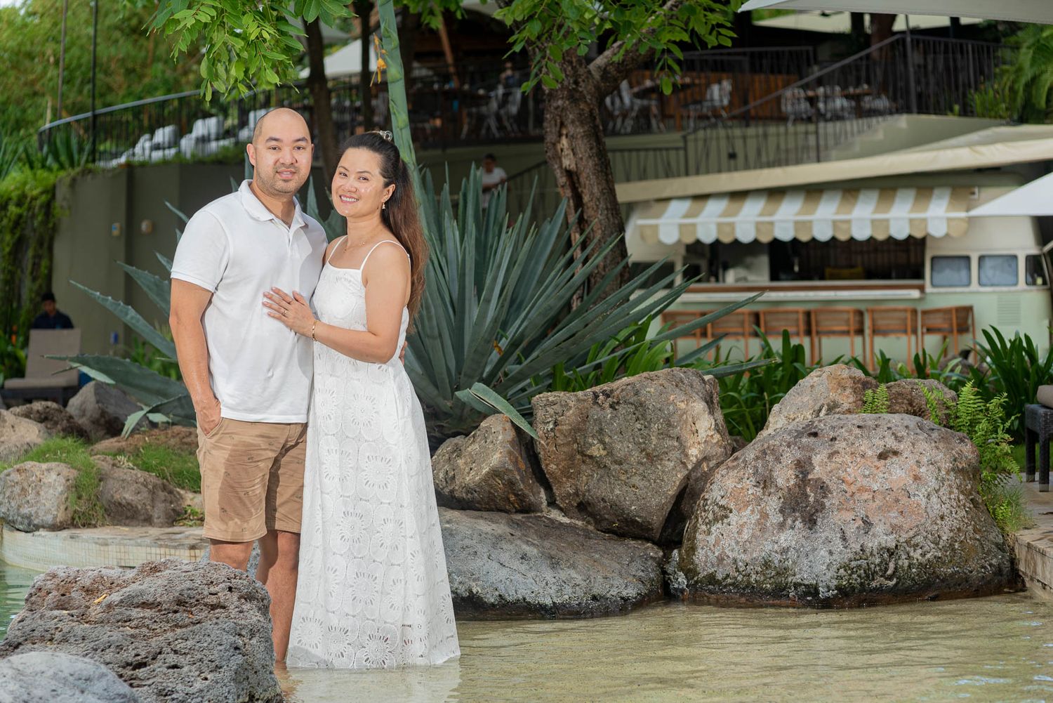 Couple posing romantically by rocky waterfront with lush tropical greenery and resort building in background.