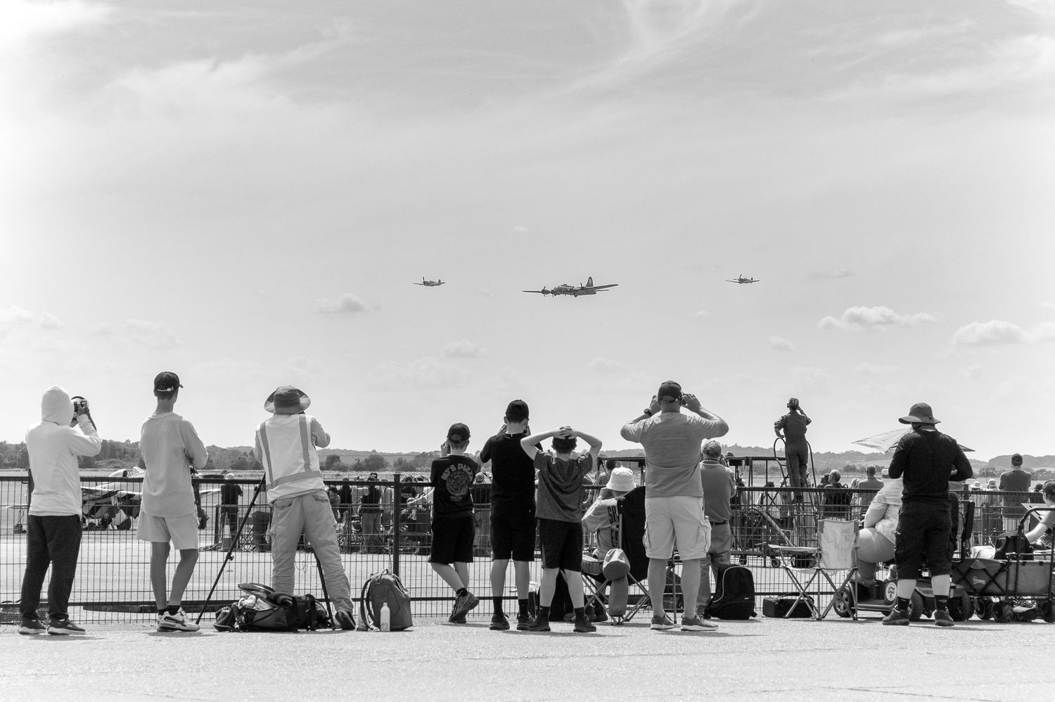 Black and white photo of spectators watching vintage aircraft fly overhead at an outdoor airshow.