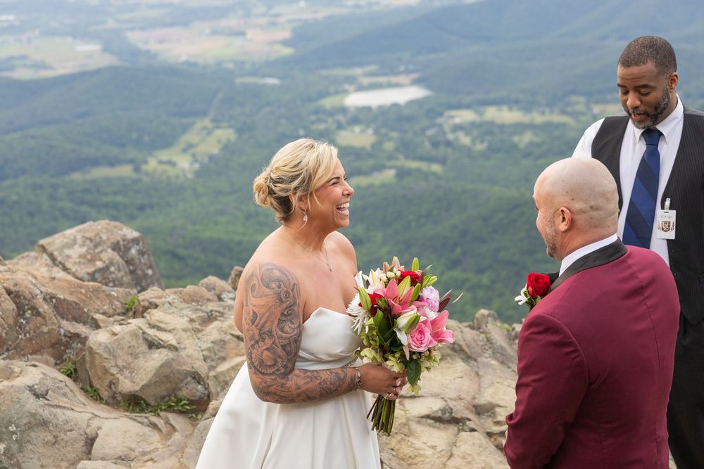 Bride and groom exchange vows on a scenic mountain summit with a officiant, surrounded by lush valley views.