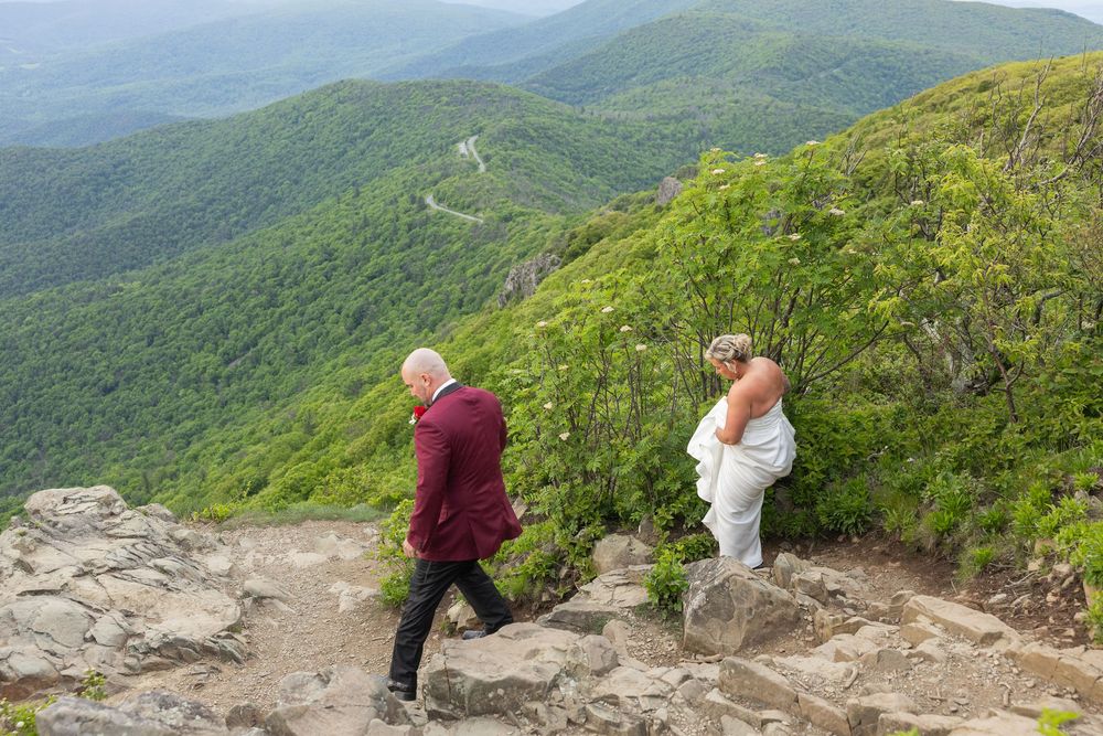 Couple in wedding attire walking on rocky mountain path with lush green valleys in background.