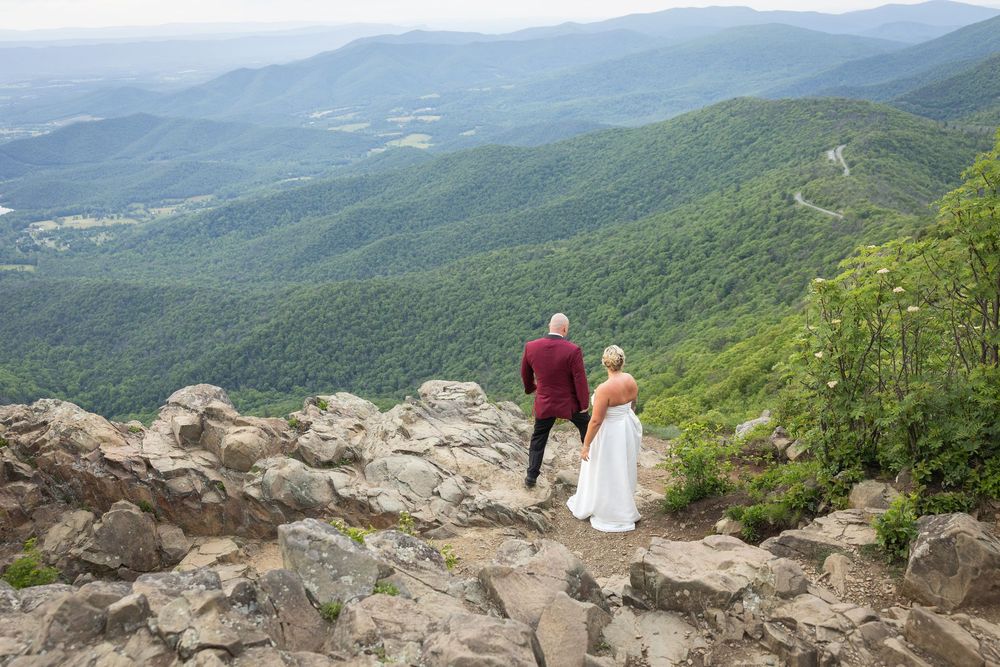 Couple in wedding attire standing on rocky overlook with panoramic green mountain valley views.