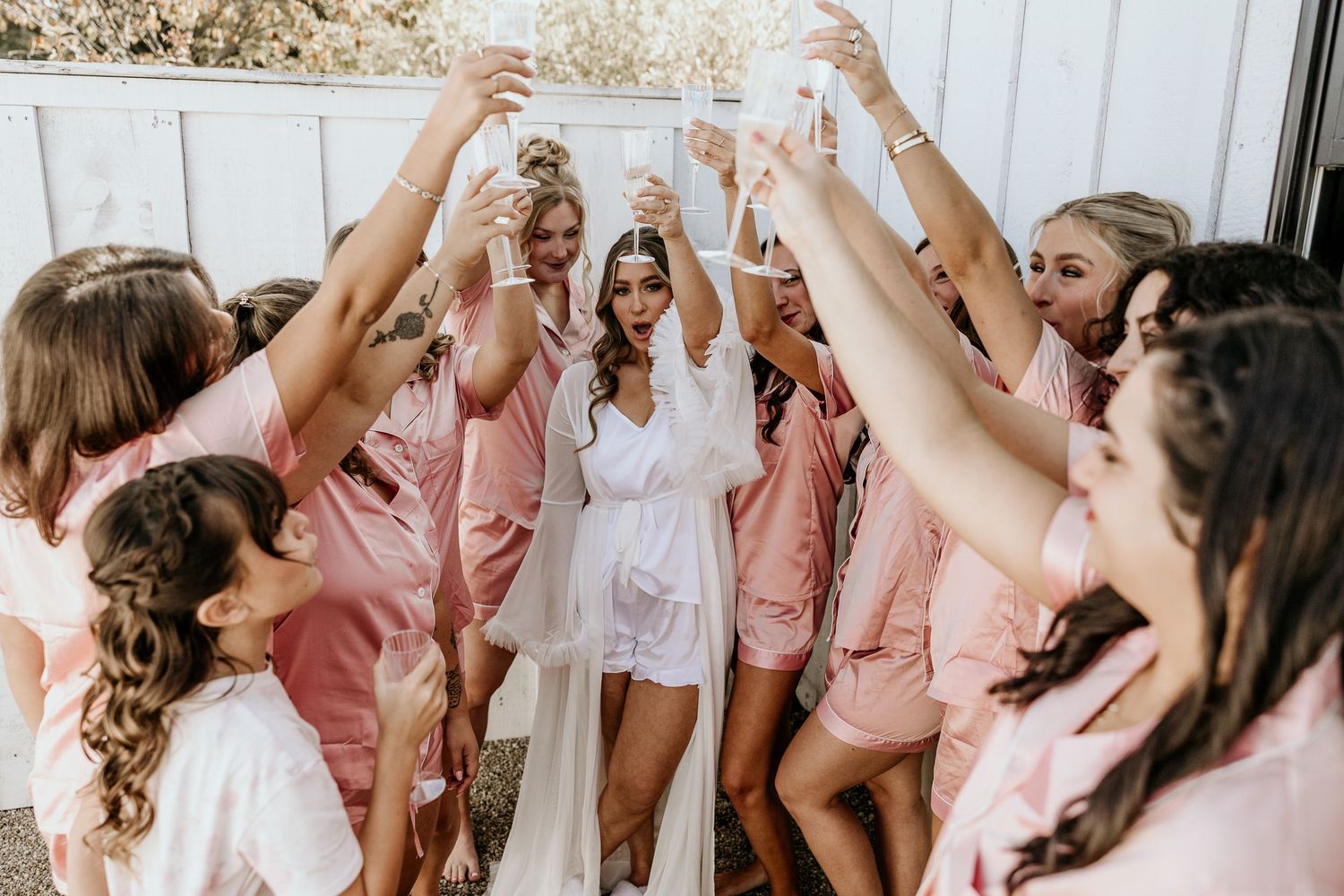 Wedding party wearing pink and white robes celebrate joyfully with raised arms in a candid group photo.