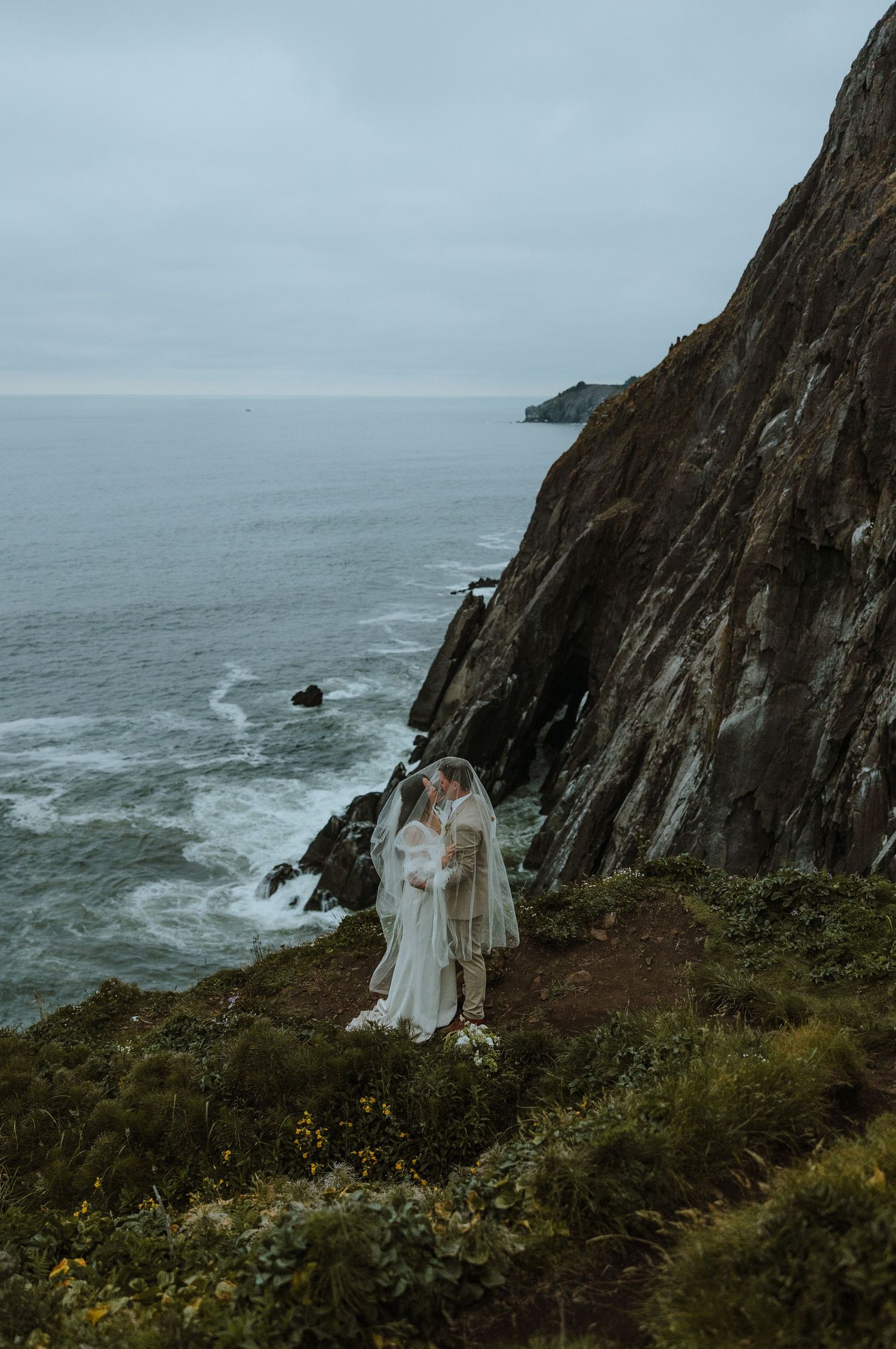 A couple in wedding attire embrace on a dramatic cliff overlooking the misty ocean coastline in moody weather.
