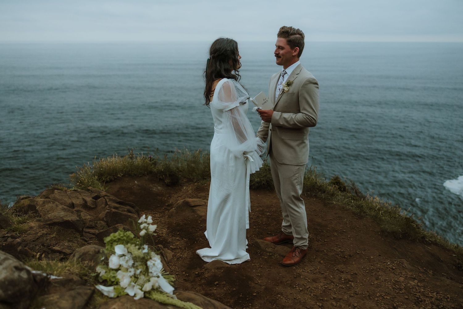 A couple in elegant wedding attire shares intimate moments on a rugged coastal cliff overlooking the misty ocean.