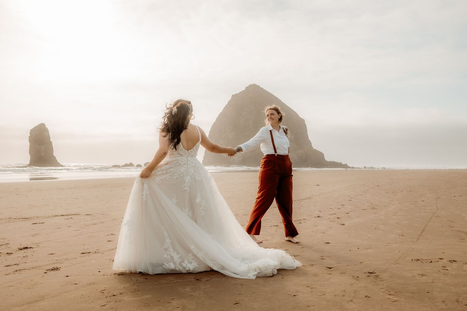 Two people in wedding attire dance together on a sandy beach at sunset with dramatic rock formations in the background.