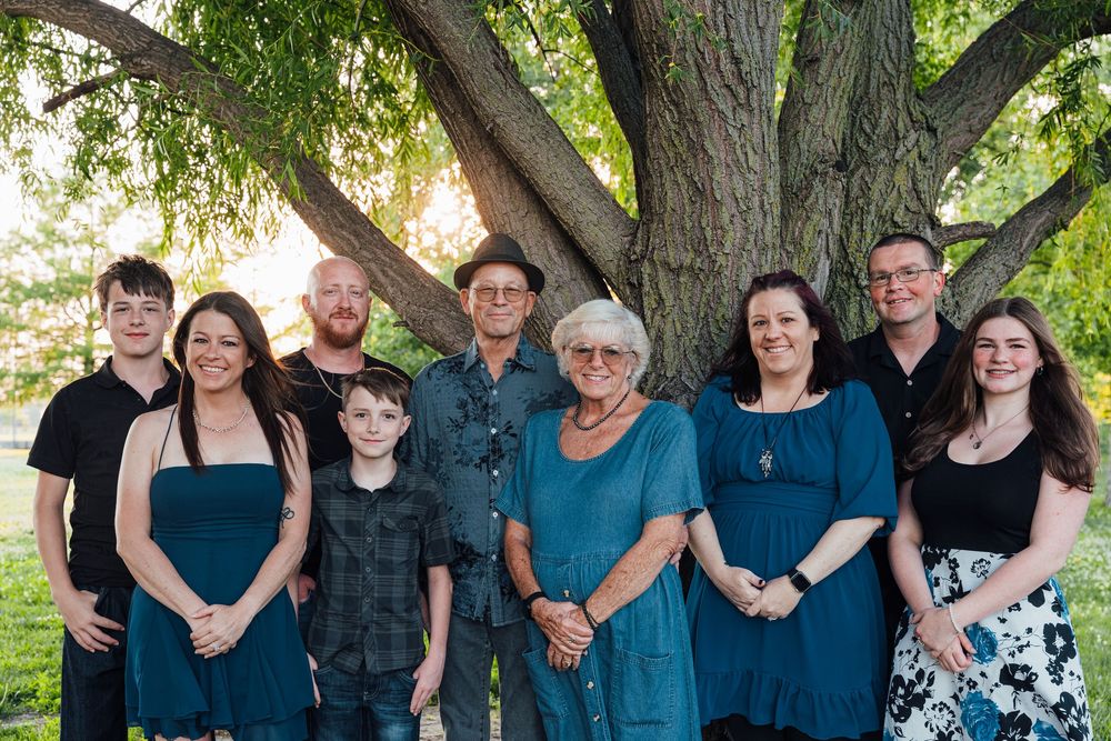 A large family group of nine people smiling together outdoors under a large tree on a sunny day.