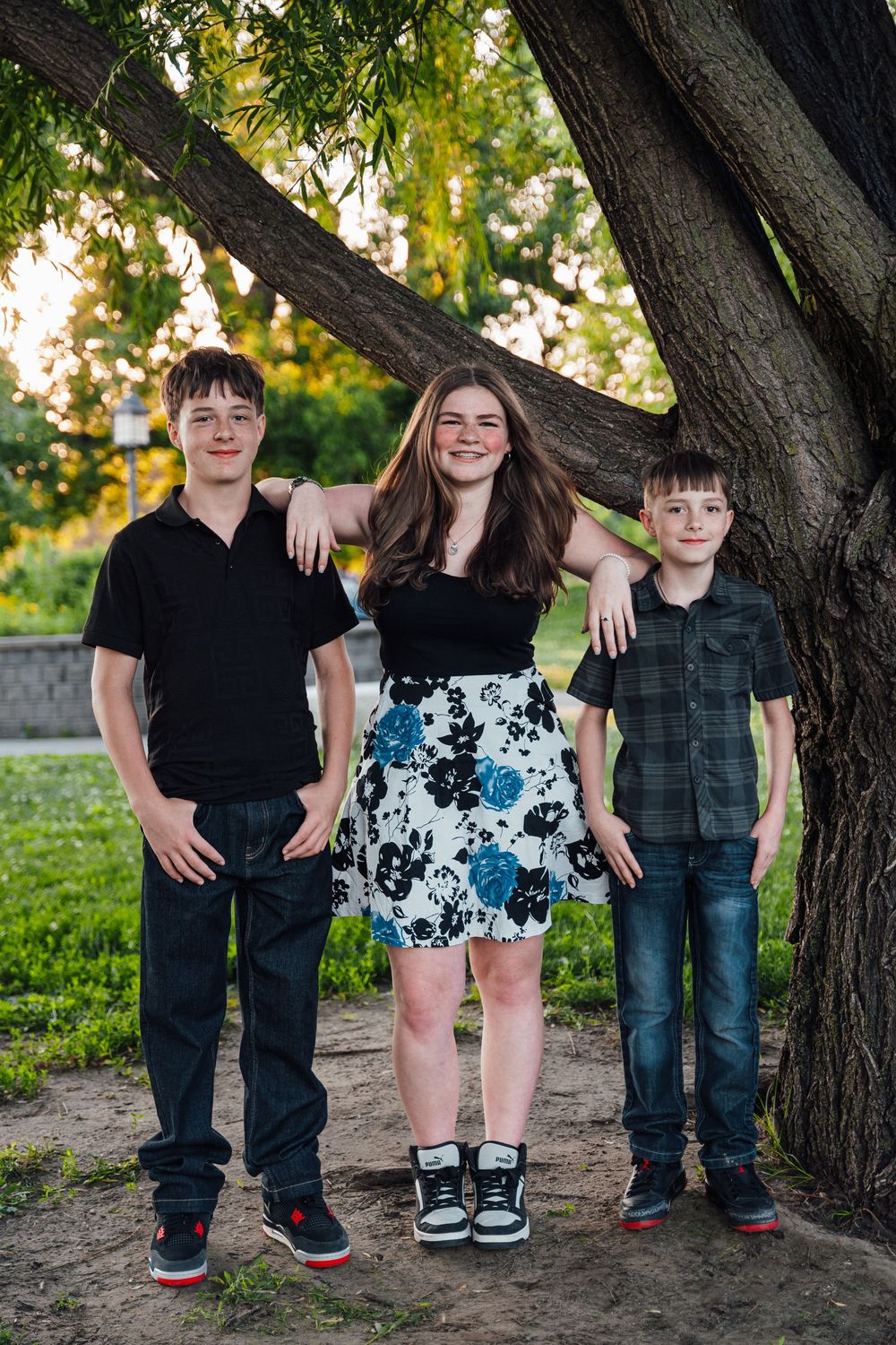 Three smiling teenagers posing under a large tree in a sunny park, wearing casual clothes including a floral skirt.