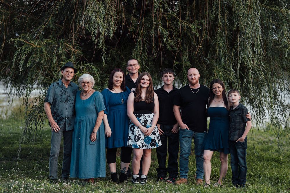 Family of nine posing outdoors under a weeping willow tree on a green lawn for a group portrait photo.