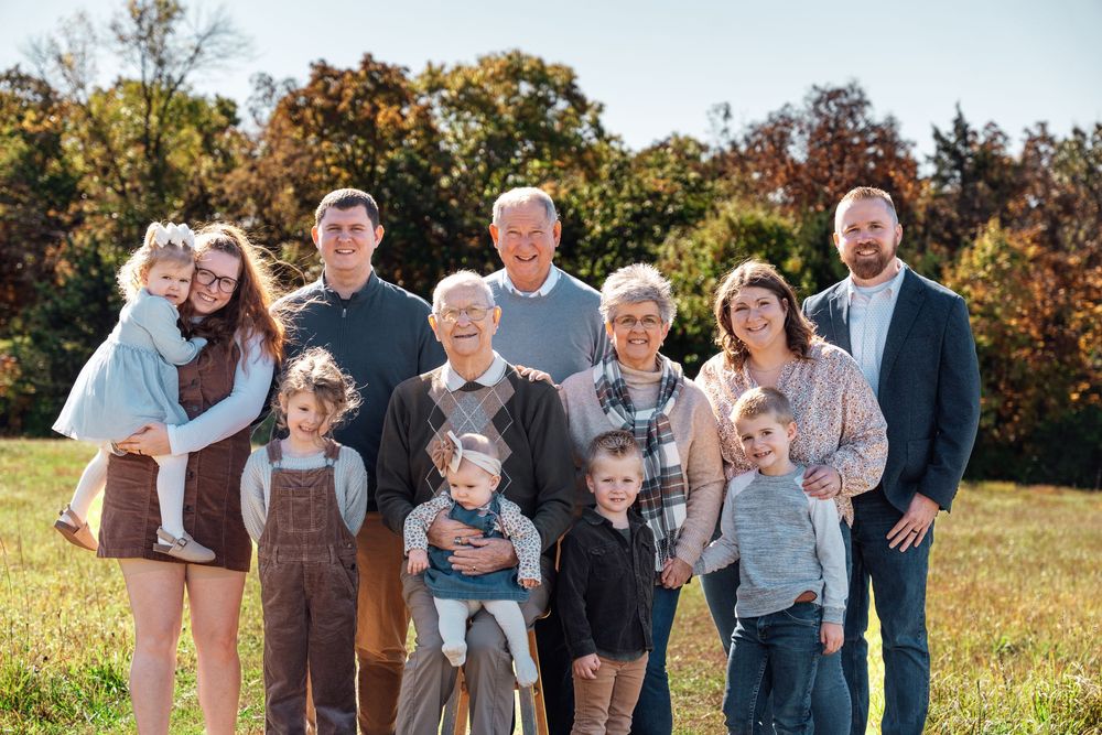 Multi-generational family portrait outdoors in autumn, smiling group of ten posed in a green field with colorful fall foliage.