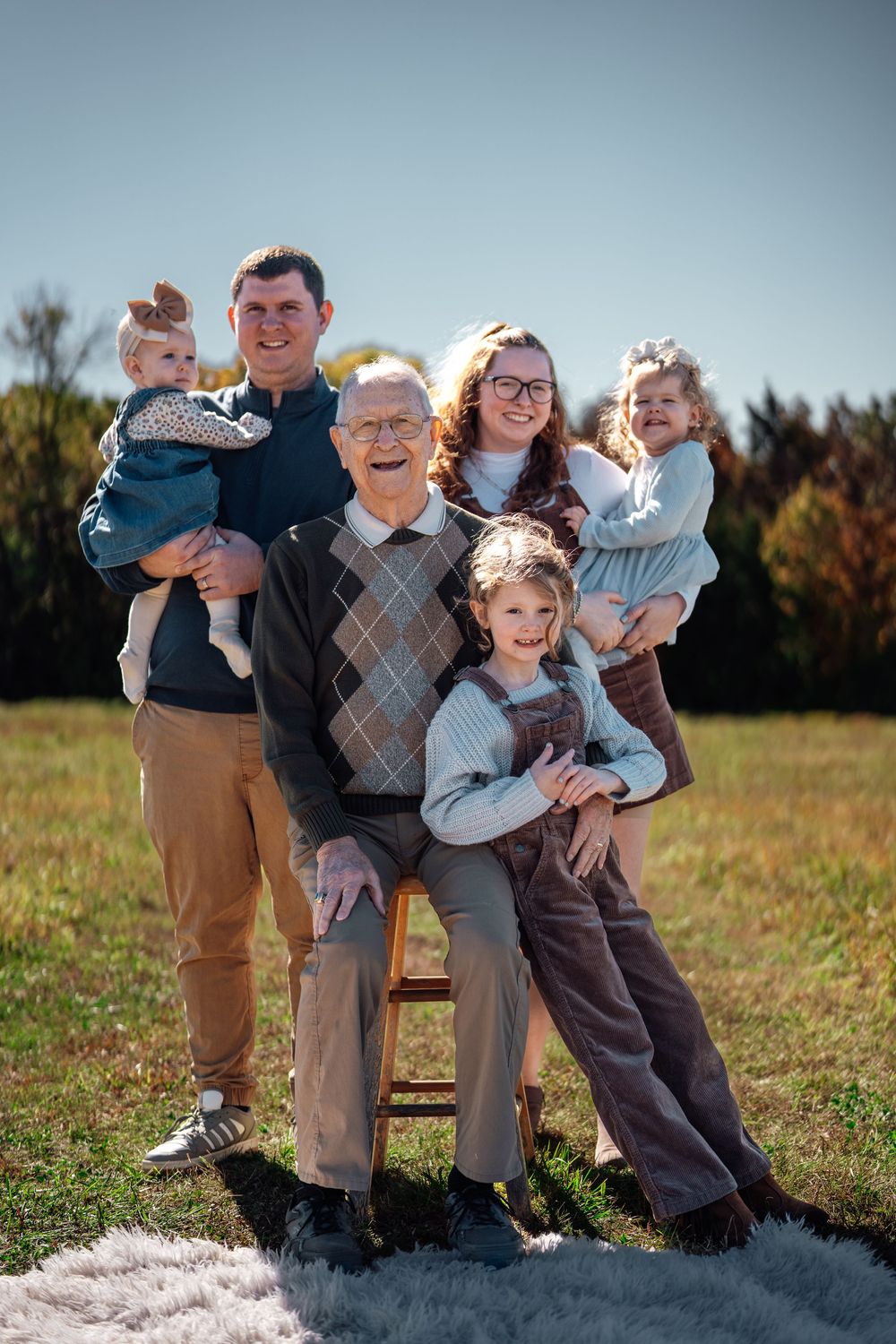 Multi-generational family portrait outdoors, elderly grandfather seated with adult couple and three young children on sunny day.