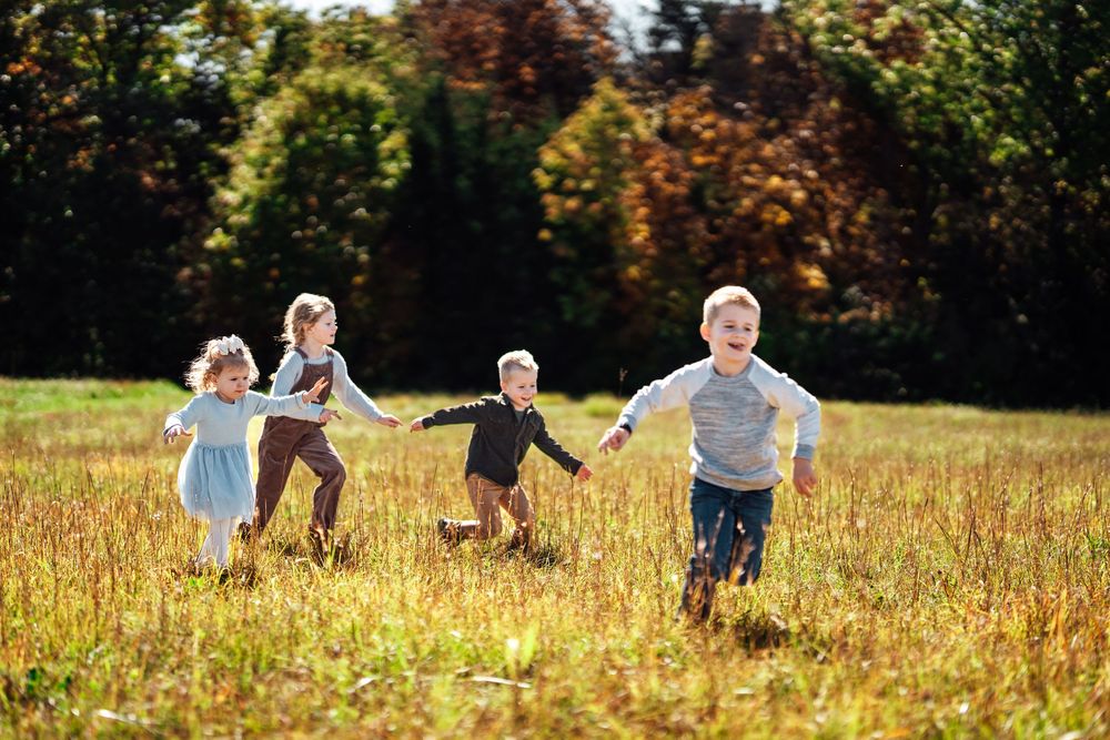 Four children running and holding hands in a sunny autumn meadow with colorful trees in the background.