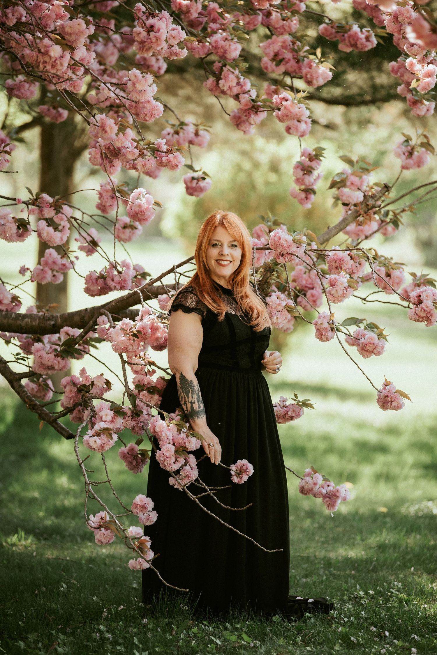 A long black dress stands beneath blooming pink cherry blossoms on a spring day in a garden setting.