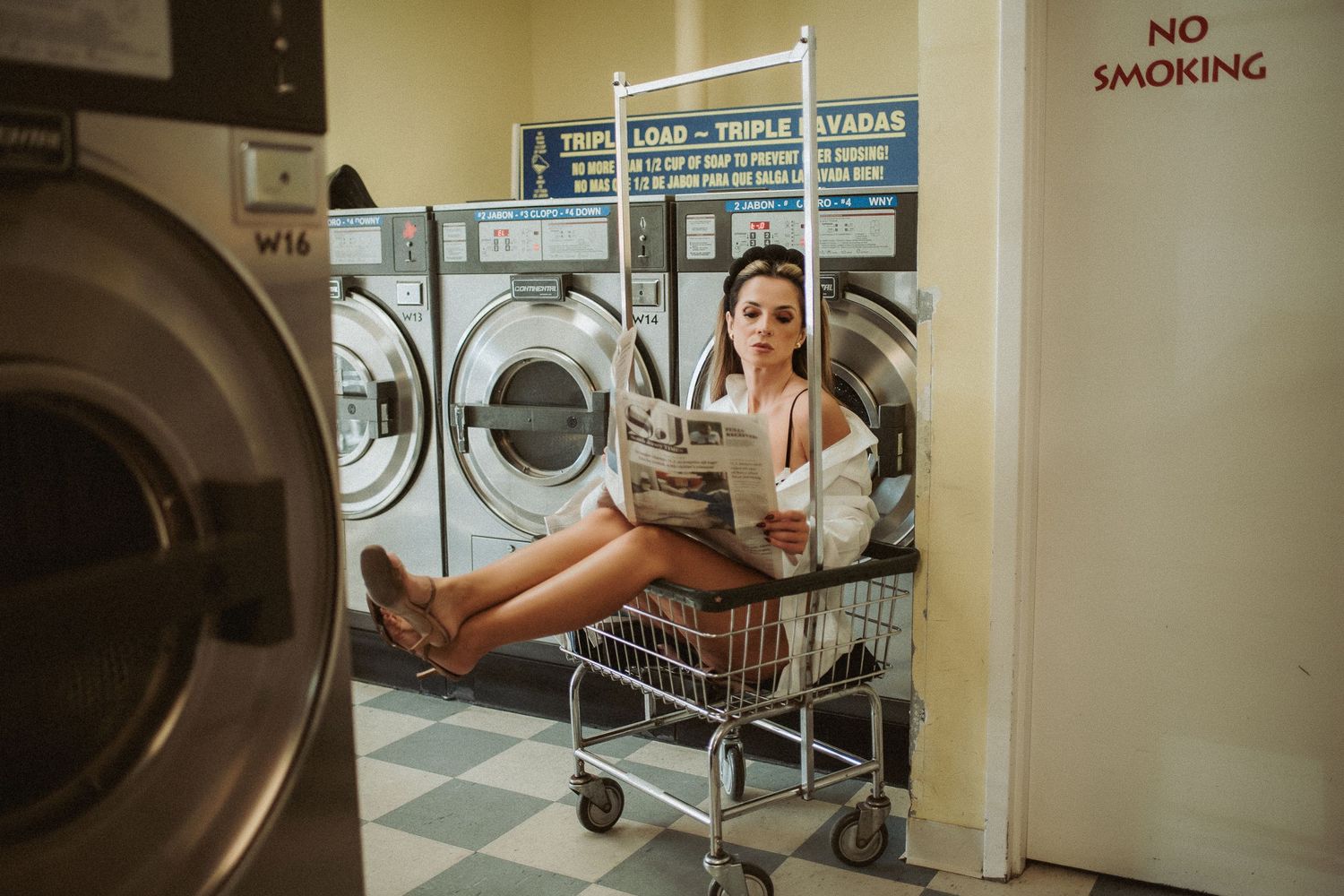 Someone sits in a laundry basket at a laundromat with washing machines and a no smoking sign visible in the background.