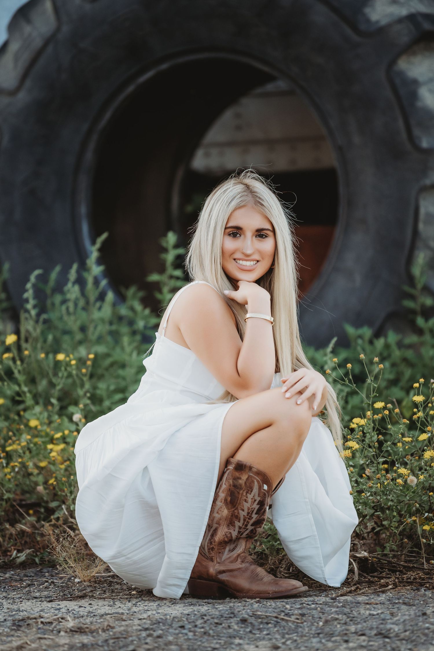 Portrait of someone in white dress and brown cowboy boots posing by large tire outdoors with wildflowers.