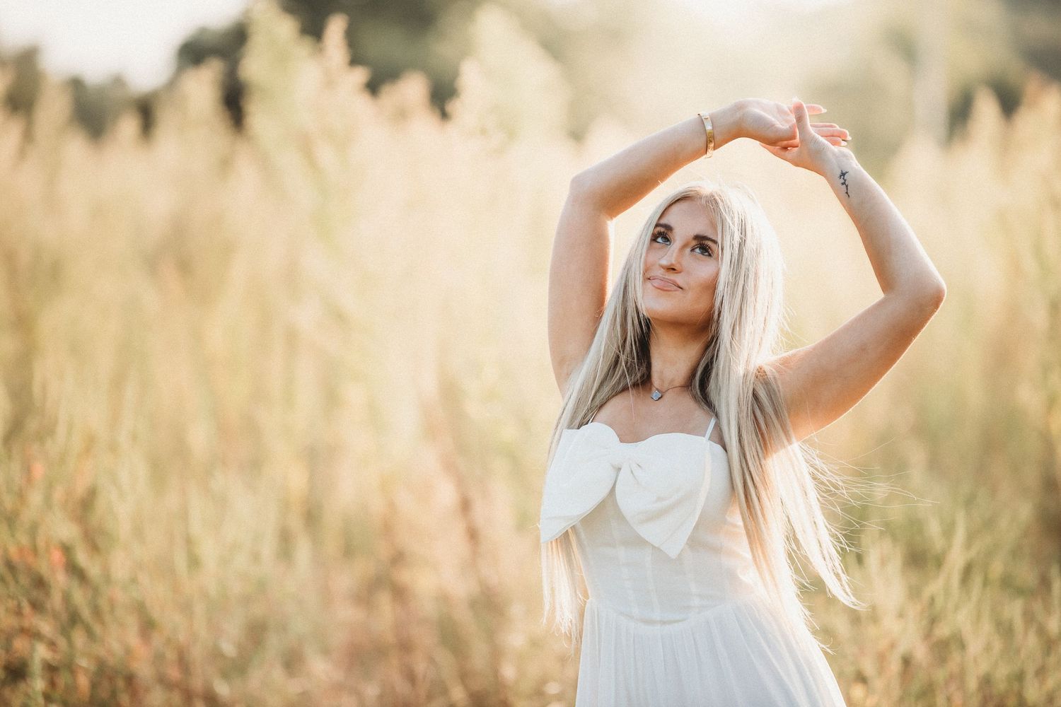 A person in a flowing white dress poses gracefully in a sunlit field of tall golden grass at sunset.