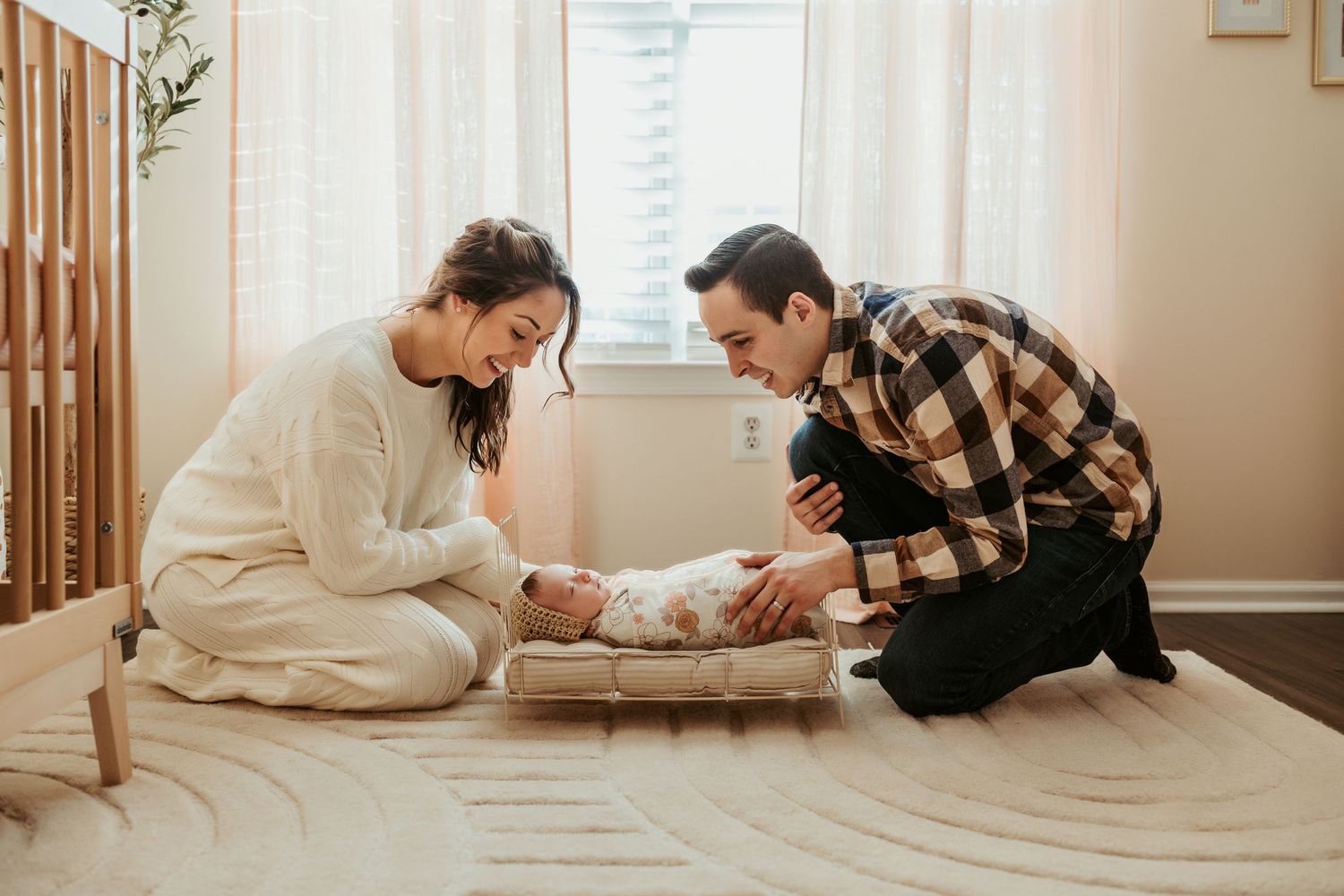Two adults kneel on a beige carpet to tend to a baby lying on a soft blanket in a bright, sunlit room.