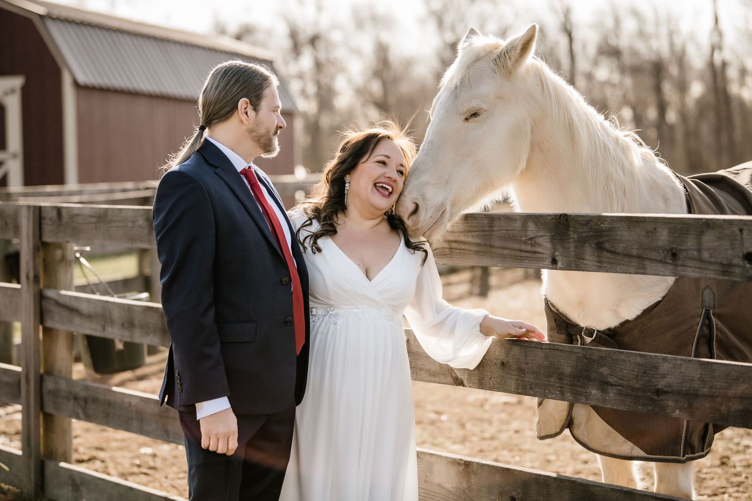 A couple in wedding attire shares a joyful moment with a white horse by a wooden fence at a rustic barn setting.