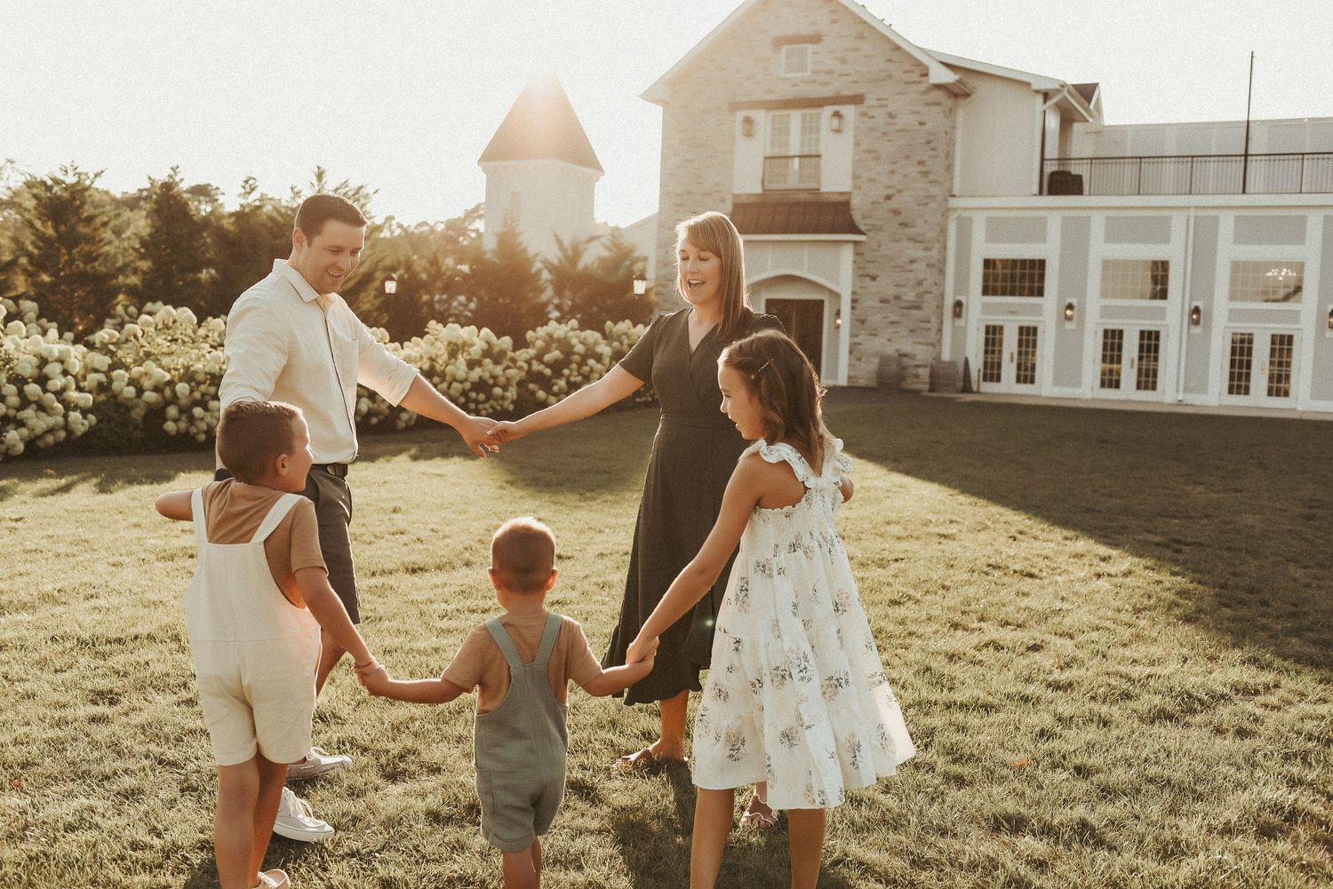 Family holding hands in circle on sunny lawn at sunset with white house and garden in background.