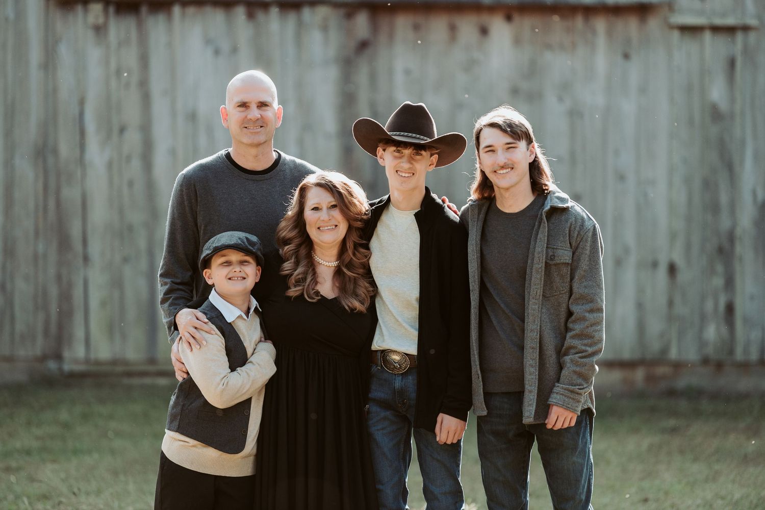 A family of five poses together outdoors against a rustic wooden barn wall on a sunny day.