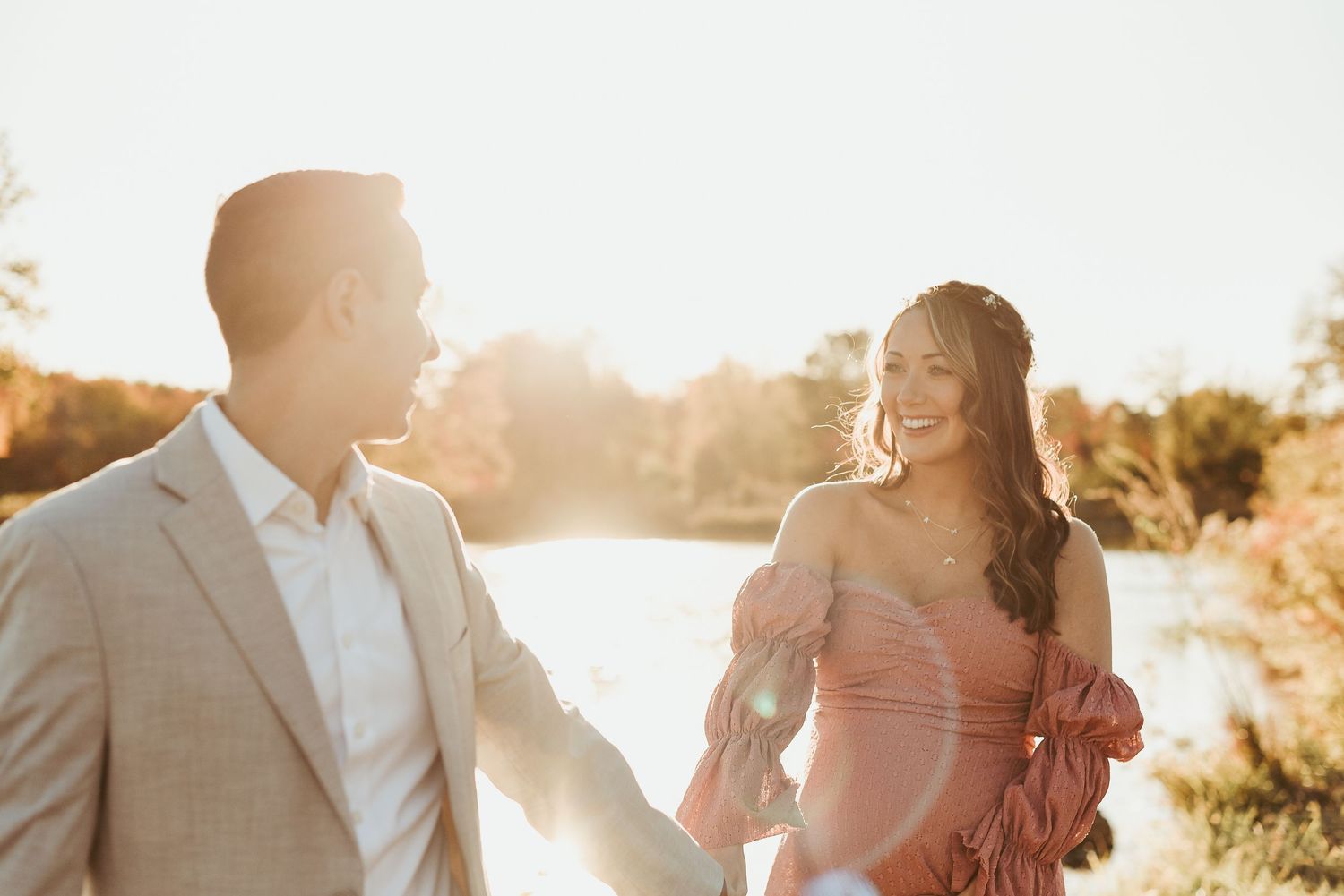 A couple holds hands and laughs together during a sunset photoshoot by a lake in golden hour lighting.