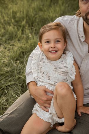 A child wearing a white lace dress sitting on grass with their adult caretaker in a summery outdoor setting.