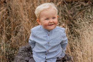 Smiling toddler wearing light blue button-down shirt sits among tall brown grass on sunny day.