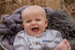 A happy baby wearing a striped shirt lies on cozy gray and purple blankets outdoors while laughing with joy.