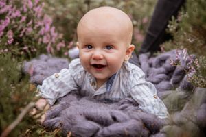A happy baby lays on a purple blanket surrounded by blooming lavender flowers outdoors.