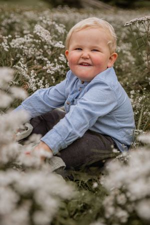 A joyful toddler in a blue shirt sits among white wildflowers on a sunny day.