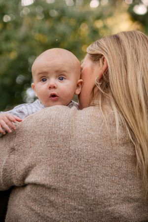 A baby with wide-eyed expression peeks over the shoulder of someone wearing a beige knit sweater outdoors.