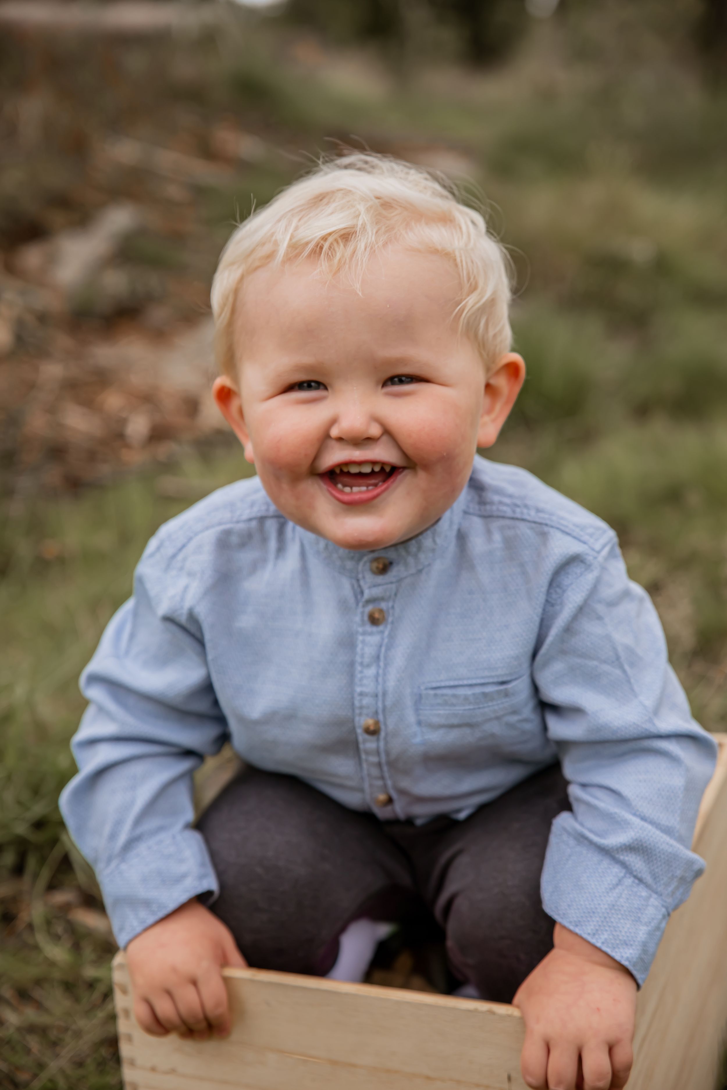 A joyful smile lights up a young child wearing a light blue button-down shirt while sitting on a wooden box outdoors.