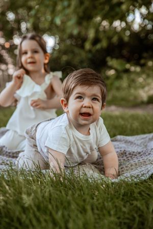 Two children playing on a blanket outdoors with joyful expressions while sitting in grass on a sunny day.