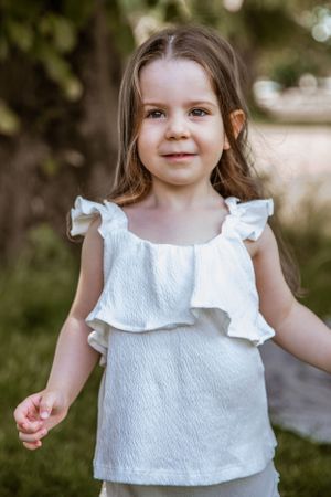 A child in a white ruffled sleeveless top smiles while standing in a garden setting.