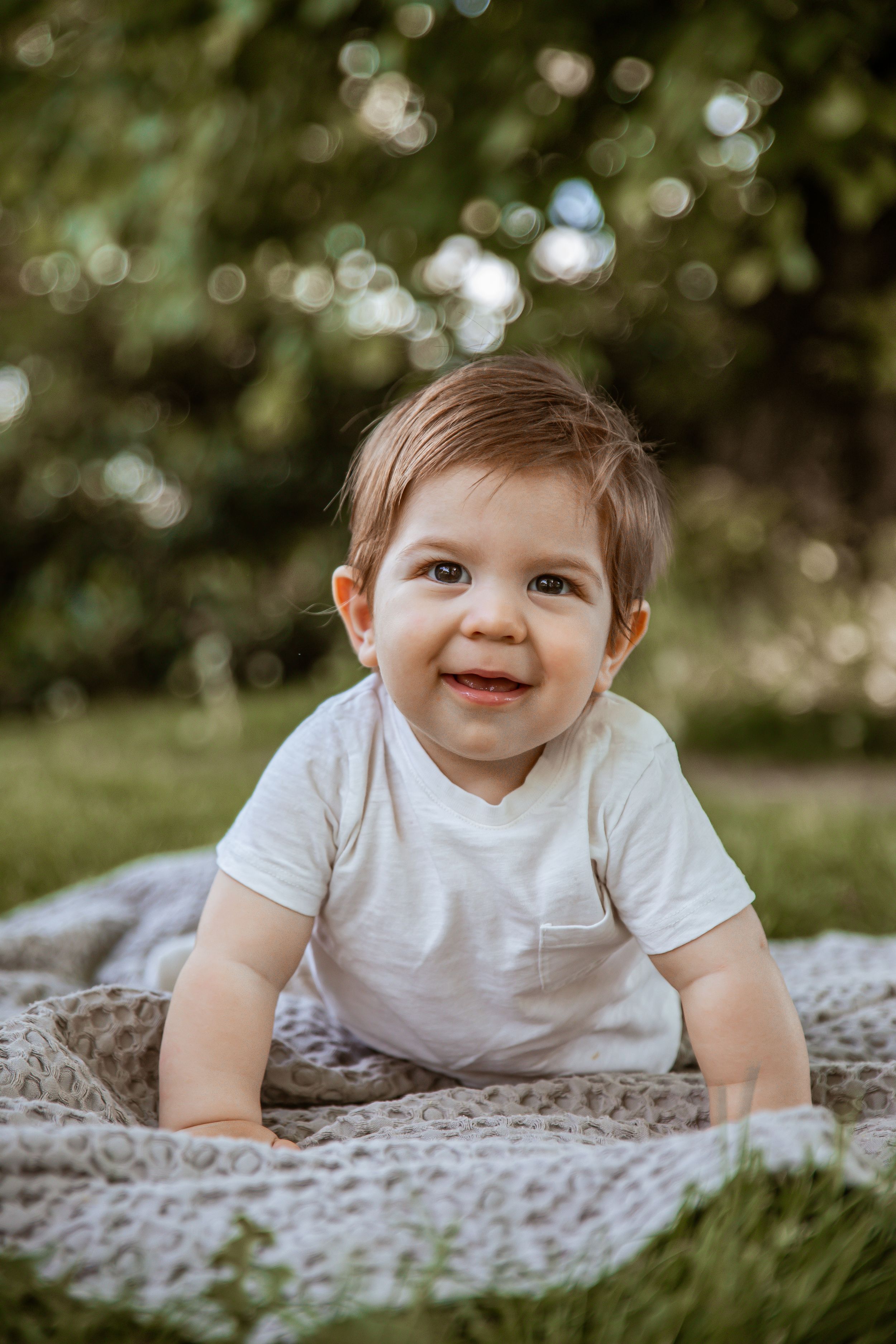 Baby in white t-shirt crawls on blanket outdoors with bokeh nature background.