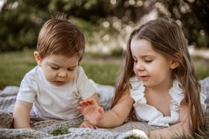 Two children sitting together on a blanket outdoors in a garden setting during a sunny day.